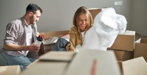 A dark haired man with a plastic sports bottle and a blonde woman sitting on the floor among carton packages