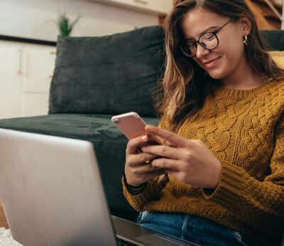 A woman texting on her phone with her laptop on her lap and back against the sofa