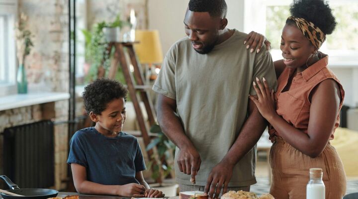 Young man cutting bread for sandwiches by table in the kitchen for his affectionate wife and their adorable little son surrounding him