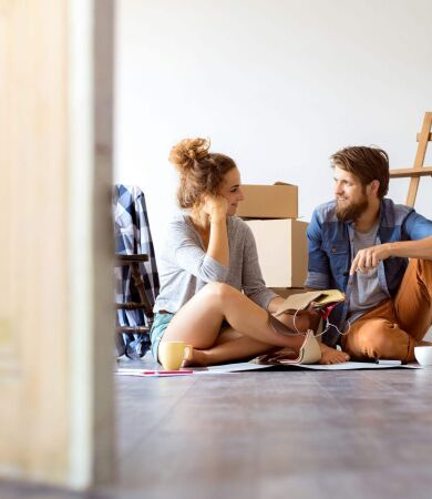 Young married couple moving into new house, sitting on the floor looking at each other with cardboard boxes behind them