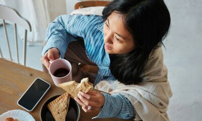 A woman drinking a coffee and eating a sandwich while wrapped up in a blanket