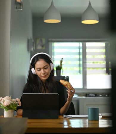 A woman on her laptop while listening to music and eating a croissant in her kitchen