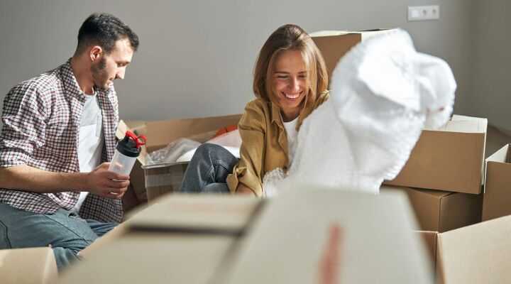 A dark haired man with a plastic sports bottle and a blonde woman sitting on the floor among carton packages