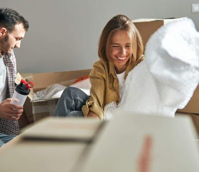 A dark haired man with a plastic sports bottle and a blonde woman sitting on the floor among carton packages