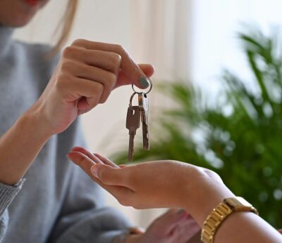 A woman wearing a grey jumping handing over a pair of keys to a woman wearing a gold watch