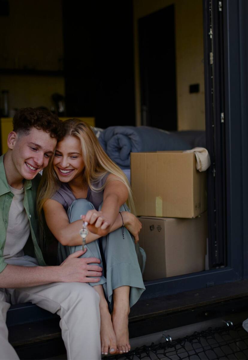 A young couple sitting outside of their new home holding the keys with moving boxes and pillows around them