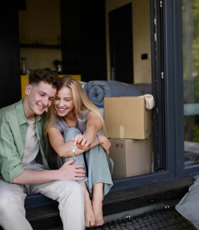 A young couple sitting outside of their new home holding the keys with moving boxes and pillows around them