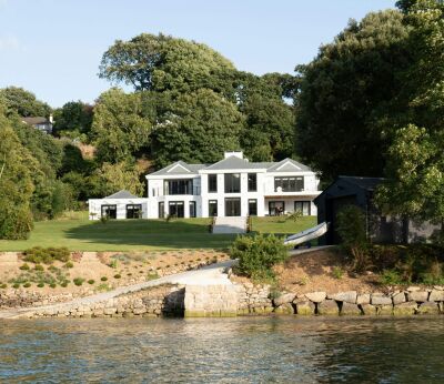 A large white house worth a million pounds surrounded by trees with a lake in front of it
