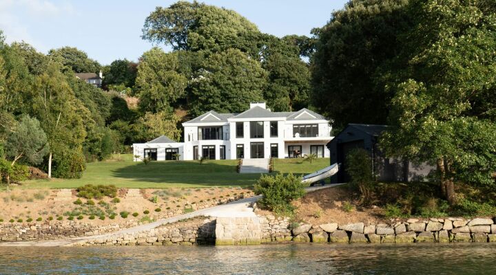 A large white house worth a million pounds surrounded by trees with a lake in front of it