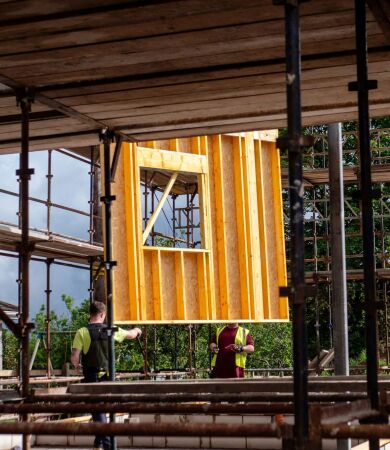 Two construction workers lifting a wooden wall frame on a scaffolding site