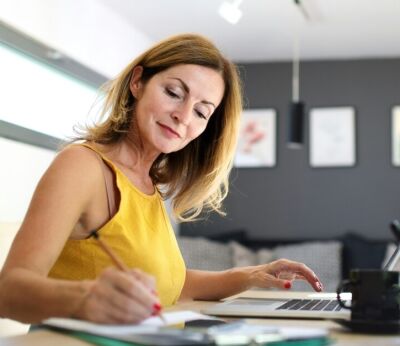 A limited company director wearing a yellow top writing in a notepad while sitting at her desk